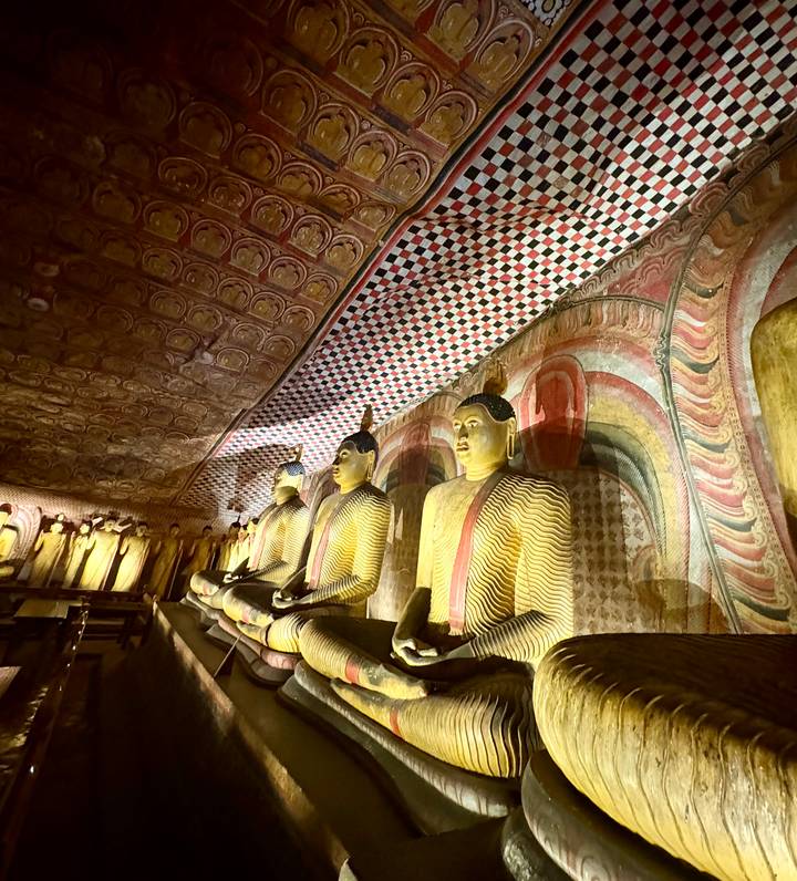 Row of golden seated Buddha statues beneath a richly painted arched ceiling inside a dim cave temple.