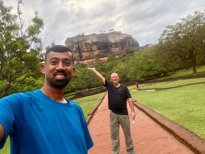 Selfie with towering Sigiriya rock fortress in the background amid lush green gardens after rain.