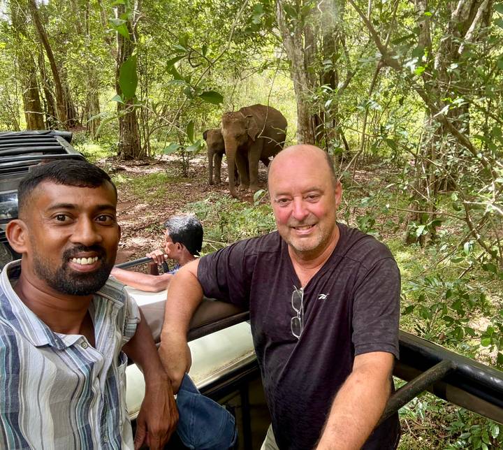 Tourist and guide smile from an open jeep while wild elephants graze in the forest behind them.