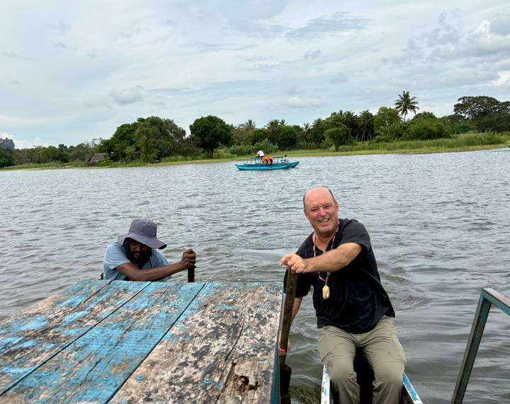 Traveler climbs from a rustic wooden raft while another man rows on a tranquil lake with palm trees beyond.