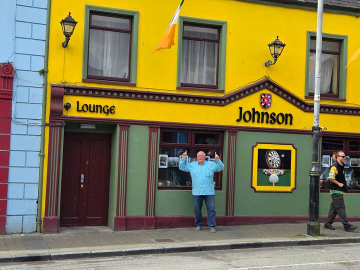 Colorful pub facade with bright yellow walls and a man posing triumphantly in front while another passer-by walks past.