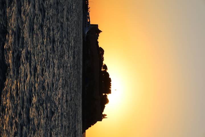 Silhouetted island fortress against a vivid orange sunset over gently rippling sea.