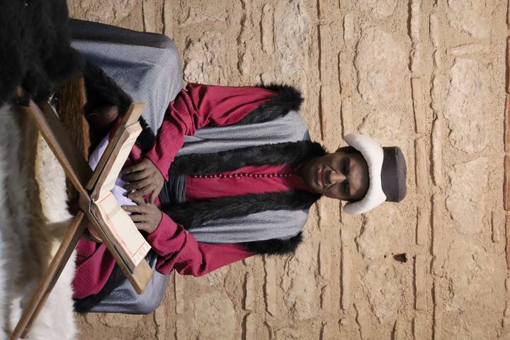 Lifelike mannequin of an Ottoman scholar in traditional robes studying an open book inside a stone-walled room.