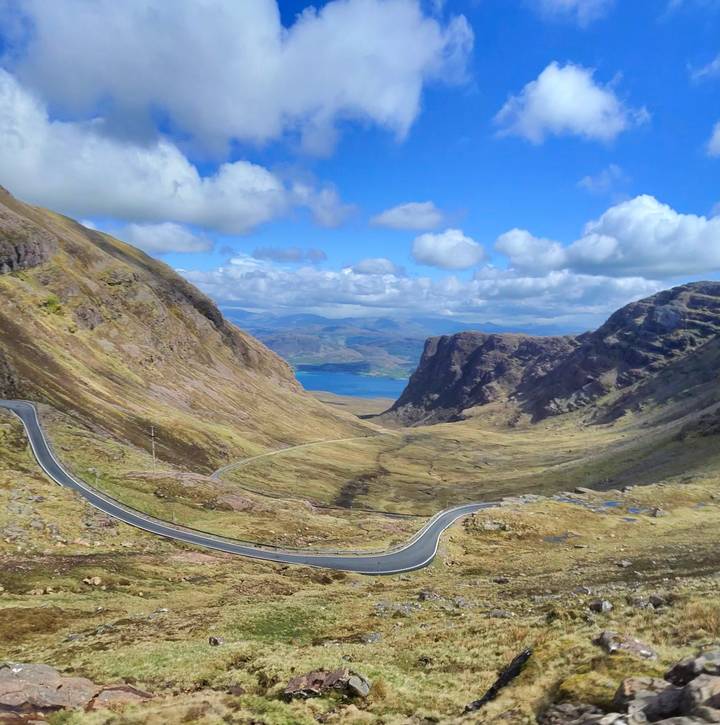 Winding mountain road descending through steep Scottish Highlands valley toward a distant blue loch.