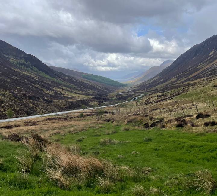 Long Highland valley carpeted with heather and crossed by a winding road under dramatic skies.