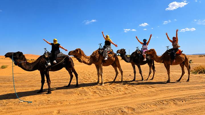 Line of tourists raising arms while mounted on camels against bright blue sky and rolling dunes.