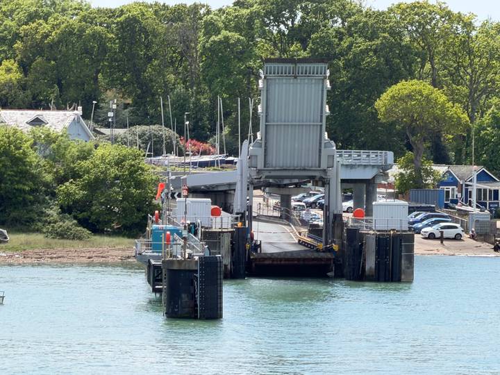Vehicle ferry ramp and terminal viewed from the water with surrounding trees and parked cars.