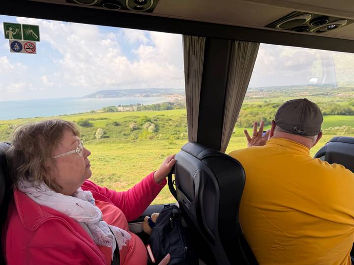 Two passengers inside a coach looking out over green coastal countryside through large windows.