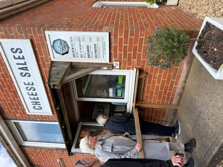 Customers buying local cheeses at a brick kiosk with large CHEESE SALES sign.