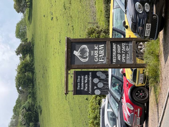 Wooden roadside sign for The Garlic Farm restaurant and shop with green hillside backdrop.