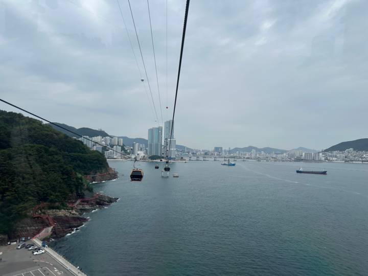 Cable cars glide above the sea toward a modern coastal skyline on a grey day in Busan.