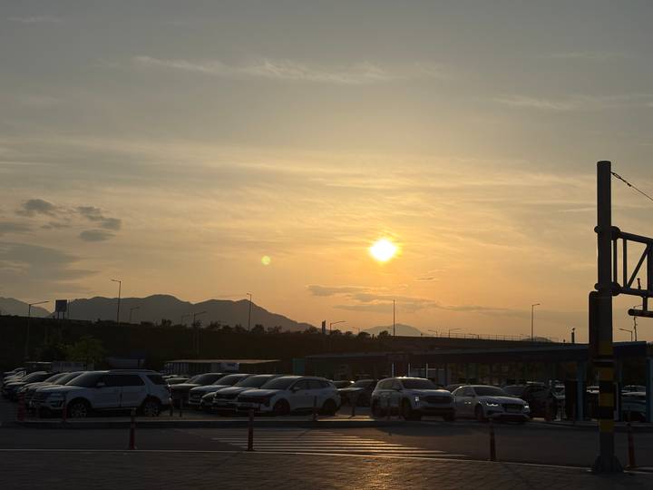 Sun sets over distant mountains beyond a car park filled with vehicles and light poles.