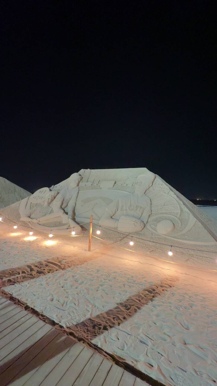 Illuminated night sand sculpture depicts baseball players under string lights.