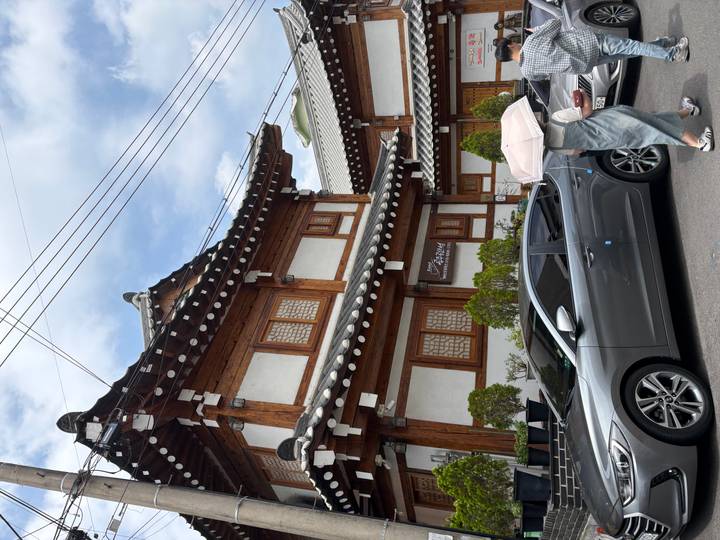 Traditional wooden hanok house stands along a street as a passerby with an umbrella walks by modern cars.