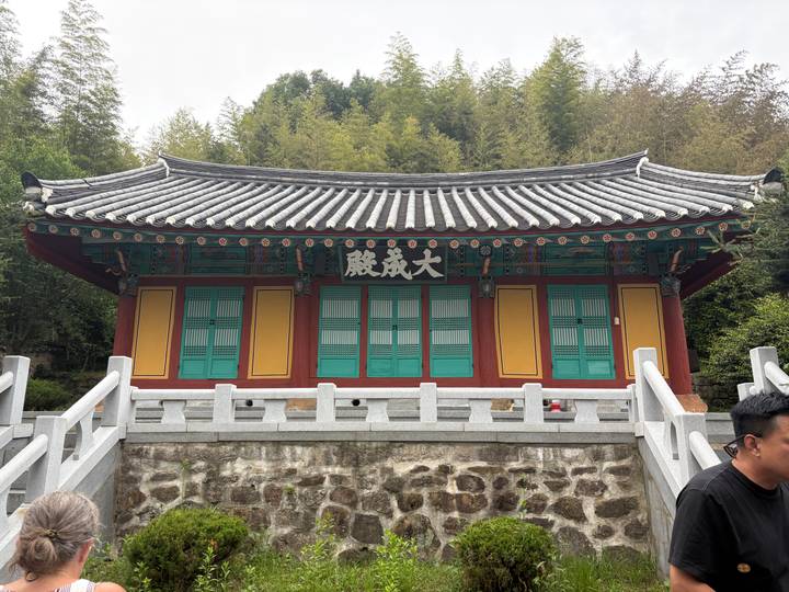 A small, colorful Buddhist shrine with teal doors and yellow walls sits at the edge of a bamboo forest.