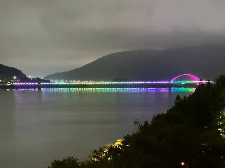 Night panorama of a lake with a long bridge lit in shifting rainbow colors and a glowing arch with mountains in the background.