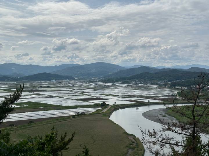 Sweeping view over flooded rice paddies, a winding river and layered mountains under a partly cloudy sky.