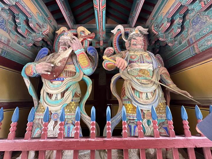 Colorful, towering wooden guardian statues inside a Korean temple gate richly painted in reds, blues and greens.