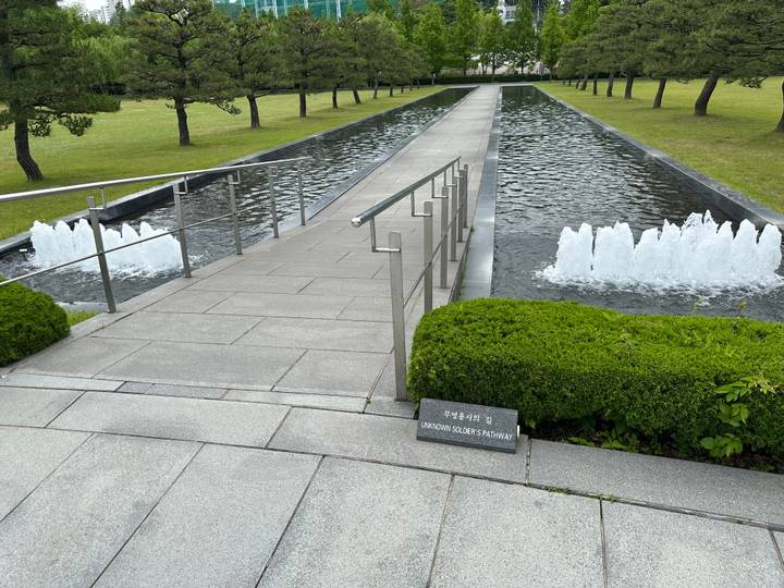 Granite walkway over a shallow reflecting pool with fountains and a plaque labelled 'Unknown Soldiers Walkway'.