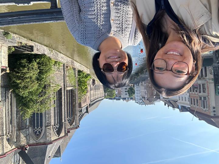 Selfie of two women on a canal bridge with historic Ghent buildings and church by the water.