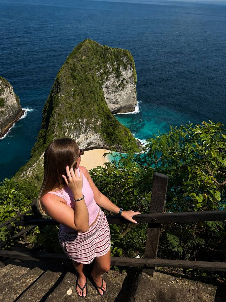 Woman sitting on viewpoint railing overlooking turquoise bay and dramatic cliffs in Nusa Penida.