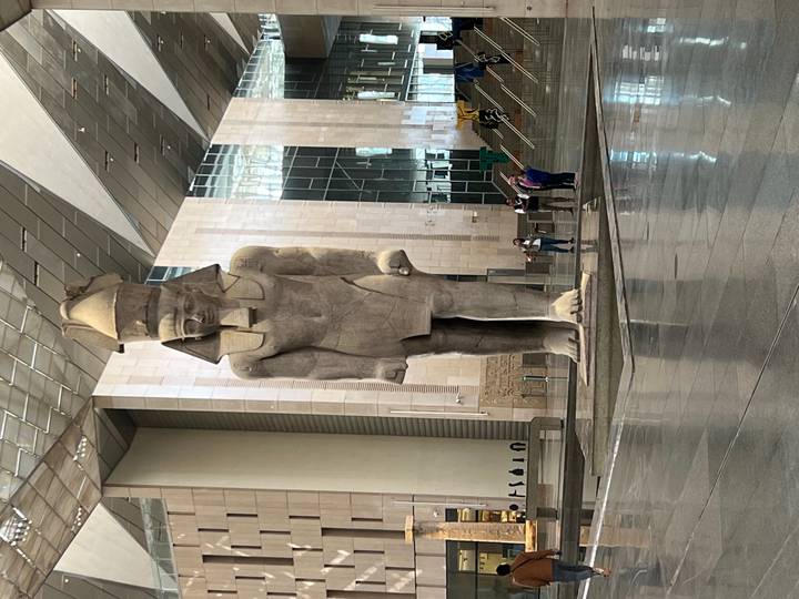 Front view of the towering Ramses II statue inside the Grand Egyptian Museum with a few visitors near its base.