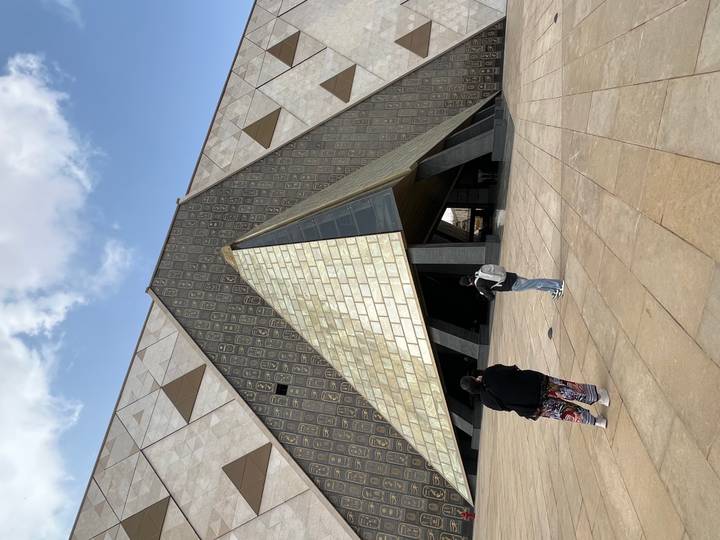 Angular pyramid-shaped entrance of the Grand Egyptian Museum with two visitors taking photos on the plaza.