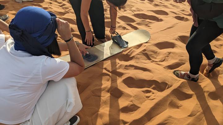 Travelers kneel on orange Sahara sand while fastening a binding to a sandboard.