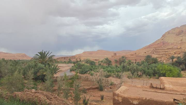 A wide view of a lush oasis valley with palms, adobe ruins and dramatic desert hills under a cloudy sky.