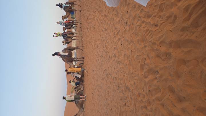 Camel riders assemble on open desert sands as the sun begins to set over the dunes.