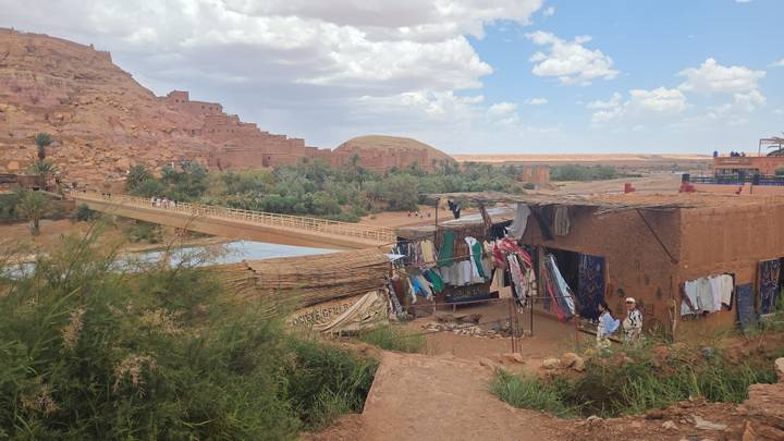 An adobe village and market stalls sit below a historic fortified hill with a bridge crossing a green river.