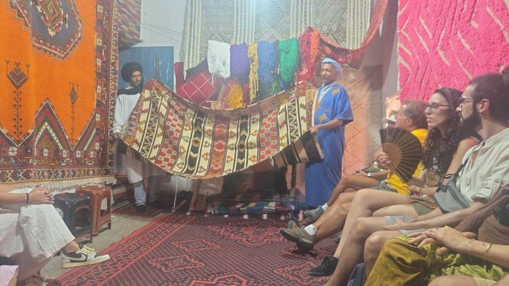 A carpet seller displays a colorful Berber rug to seated travelers inside a textile shop.