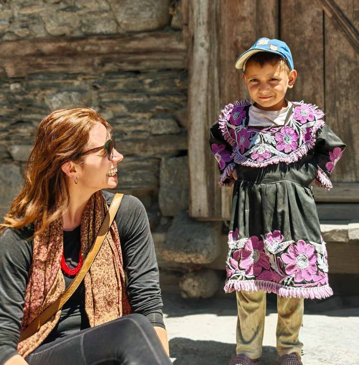 A smiling traveler chats with a young girl in an embroidered dress outside a stone building.