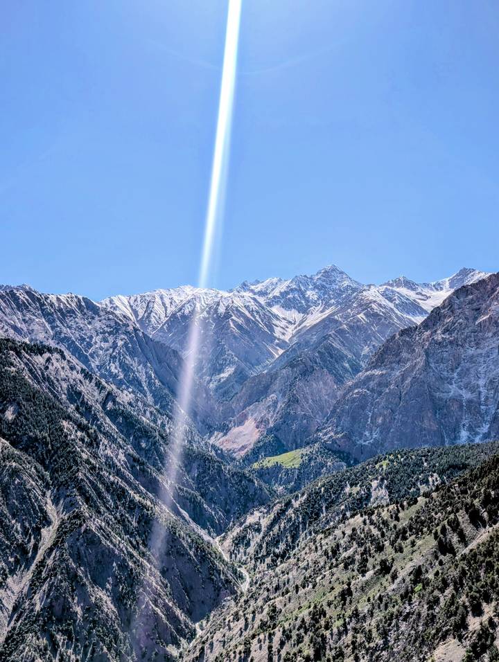 Snow-capped peaks rise behind forested slopes with a dramatic vertical sunbeam cutting through the clear blue sky.