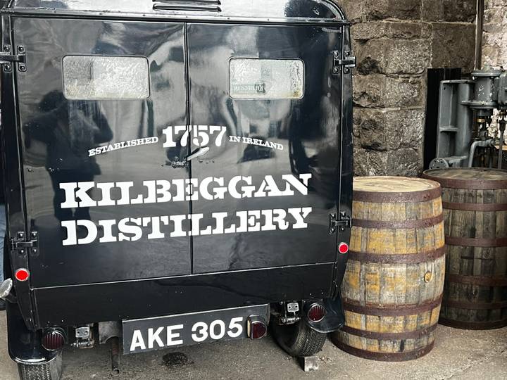 Vintage black delivery van with ‘Kilbeggan Distillery’ branding parked beside barrels