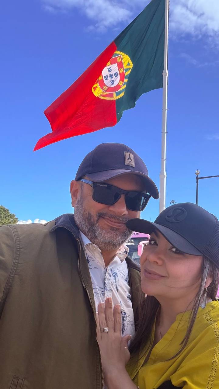 Close-up selfie of a couple wearing caps with blue sky and red flag behind them