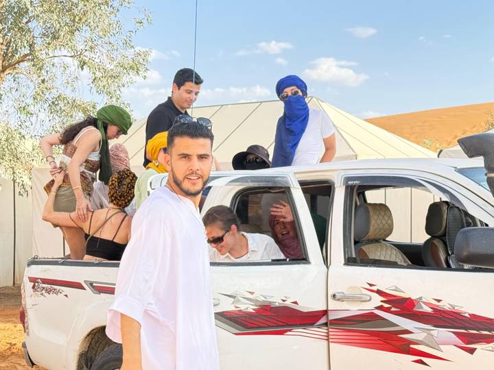 Travelers laugh and pose around a white pickup truck beside desert tents under a bright sky.