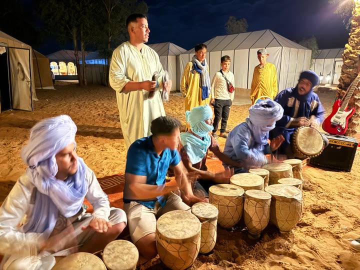 Night-time drum circle with performers in traditional Tuareg attire playing hand drums at a desert camp.