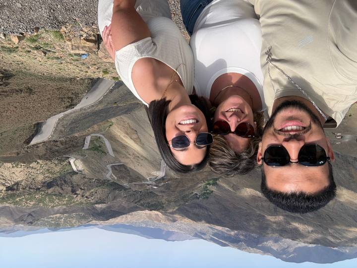 Selfie of three smiling travellers with winding mountain road and rugged Atlas Mountains in the background.
