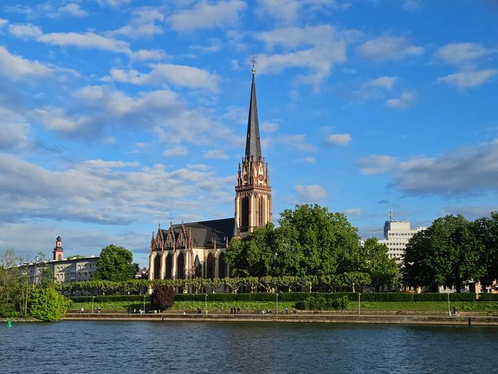 Neo-Gothic church spire rising above leafy riverside park under blue sky in Frankfurt.