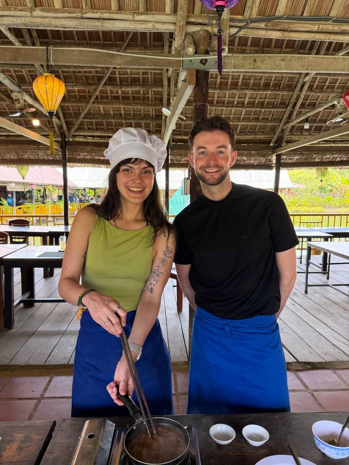 Smiling couple in cooking uniforms preparing food inside a rustic open-air kitchen.