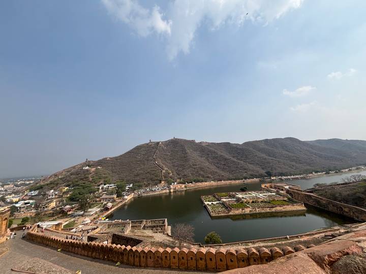 Elevated view of Maota Lake and the rugged hillside walls leading up to a fort complex, with the city of Jaipur sprawling to the left under a clear blue sky.
