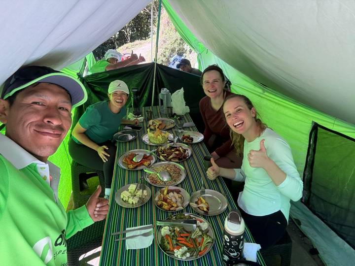 Happy trekkers sit inside a bright green mess tent sharing an abundant camping meal and giving thumbs up.
