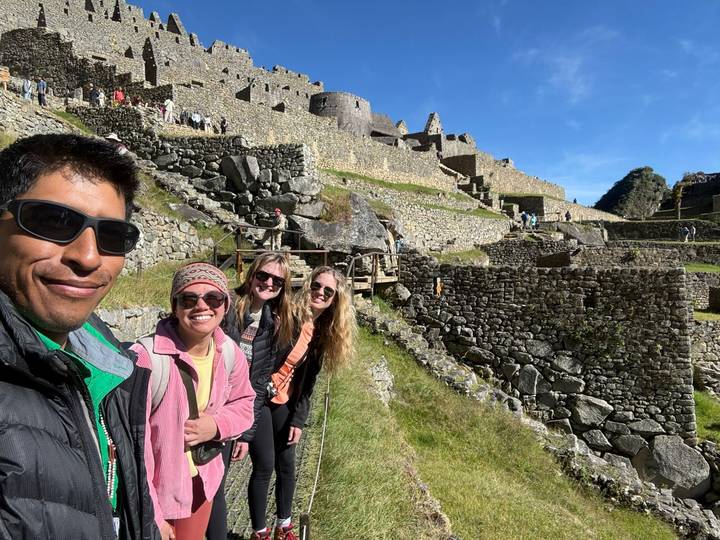 Smiling trekkers stand on stone terraces of Machu Picchu with ruins rising behind them.