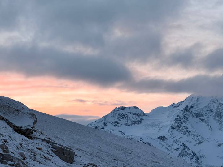 Soft pink sunrise light bathes snow-capped Himalayan peaks beneath dramatic clouds.