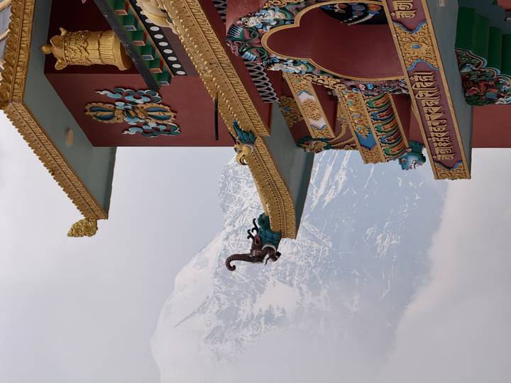 Ornate Tibetan-style monastery eaves decorated with vibrant carvings, backed by a snowy peak.