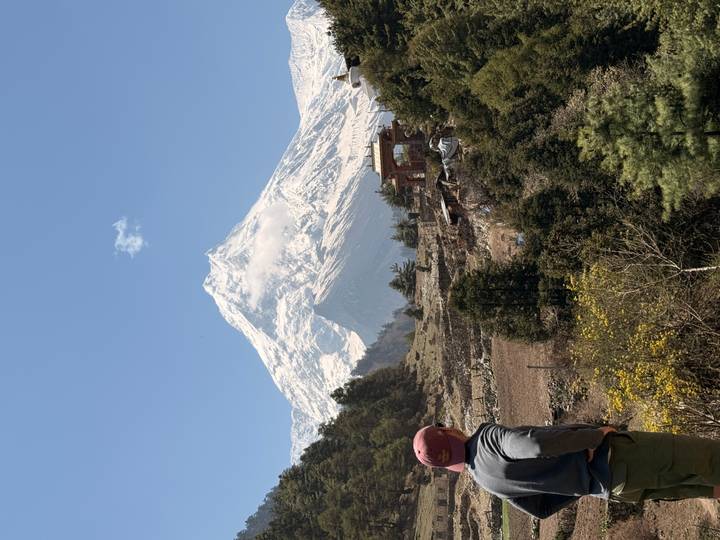 A hiker gazes at a towering snow-clad mountain rising above a rural Nepali valley with a small gate.