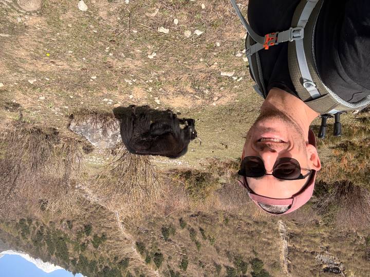 Smiling trekker selfies with a grazing yak on an alpine meadow trail.