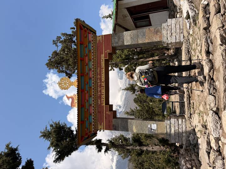 Hikers pass beneath a decorative arch reading 'Lotus Feet of Manaslu Heritage' on a forested trail.