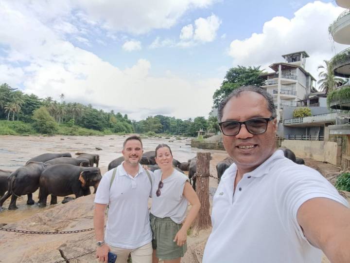 Selfie of three travellers by a river where a herd of elephants bathes behind them.